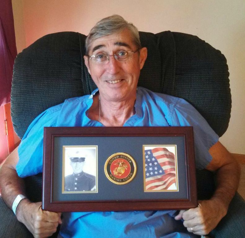 Man sitting in chair holds frame with black and white photo of self, Marine Corps emblem, and American flag image